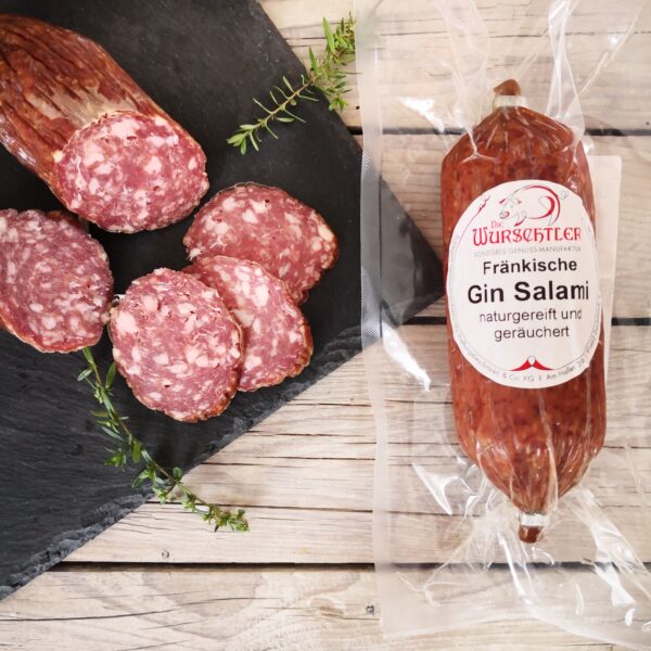 Salami mit Gin am Stück und aufgeschnitten angerichtet auf einer Schieferplatte und garniert mit Kräutern; HofladenBOX